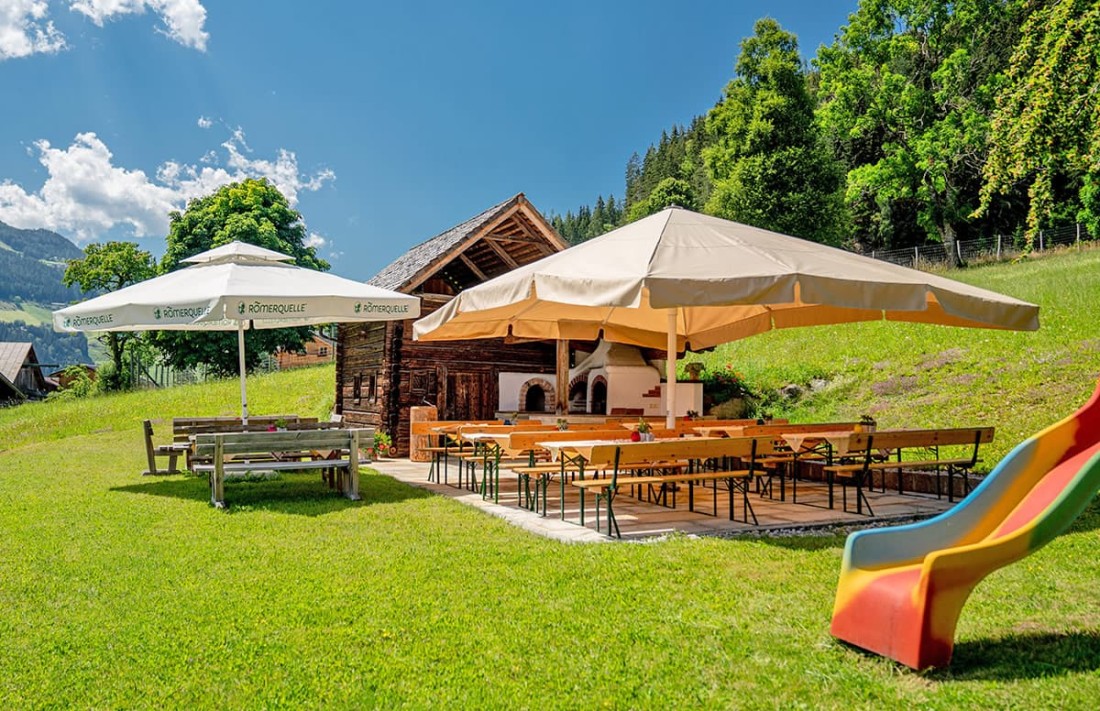 Rustikale Terrasse mit Holztischen, Sonnenschirmen und Rutsche vor einer Holzh&uuml;tte